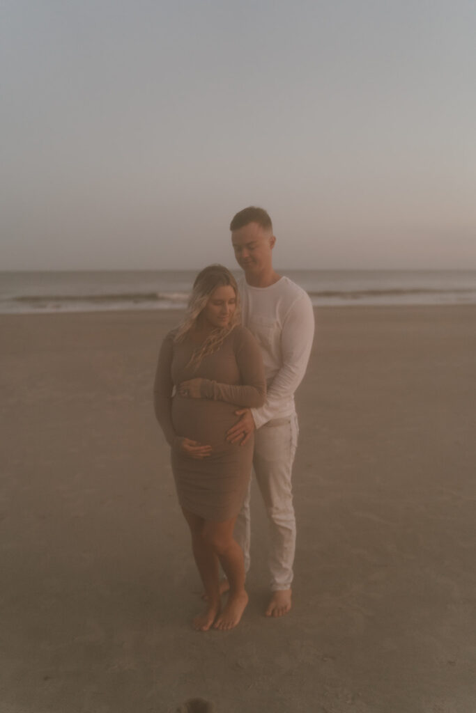 Couple walking on the beach holding baby bump during maternity shoot 