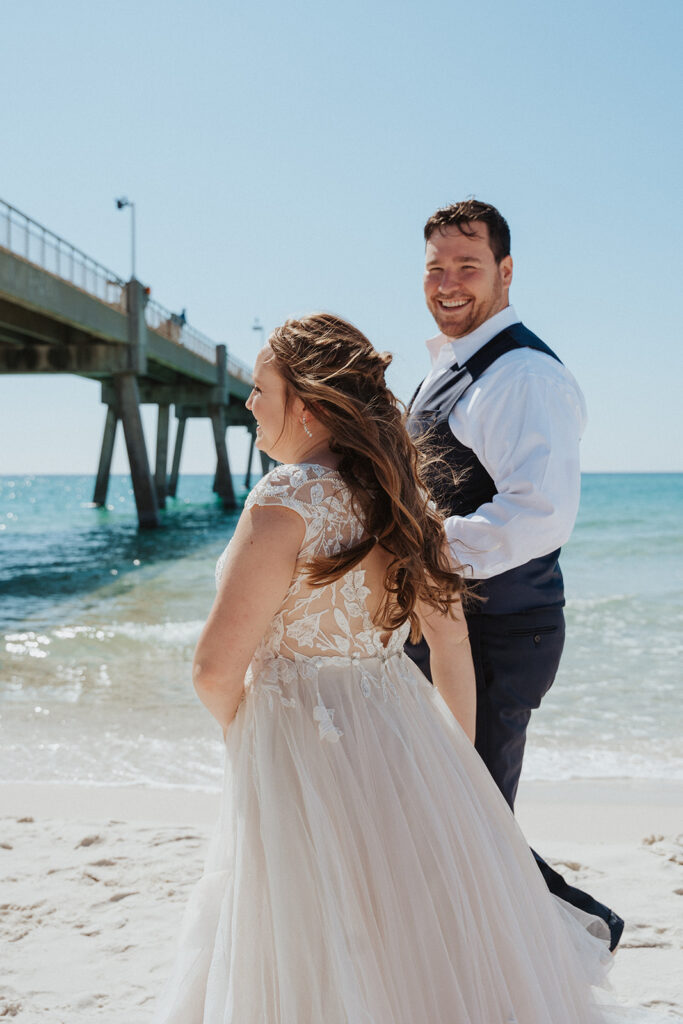 groom smiling while walking on beach with bride