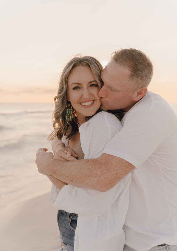 A man hugs a smiling woman from behind and kisses her cheek on a beach at sunset. Both wear white shirts, and she has wavy hair and statement earrings. The mood is warm and affectionate—perfect inspiration for seaside family pictures.