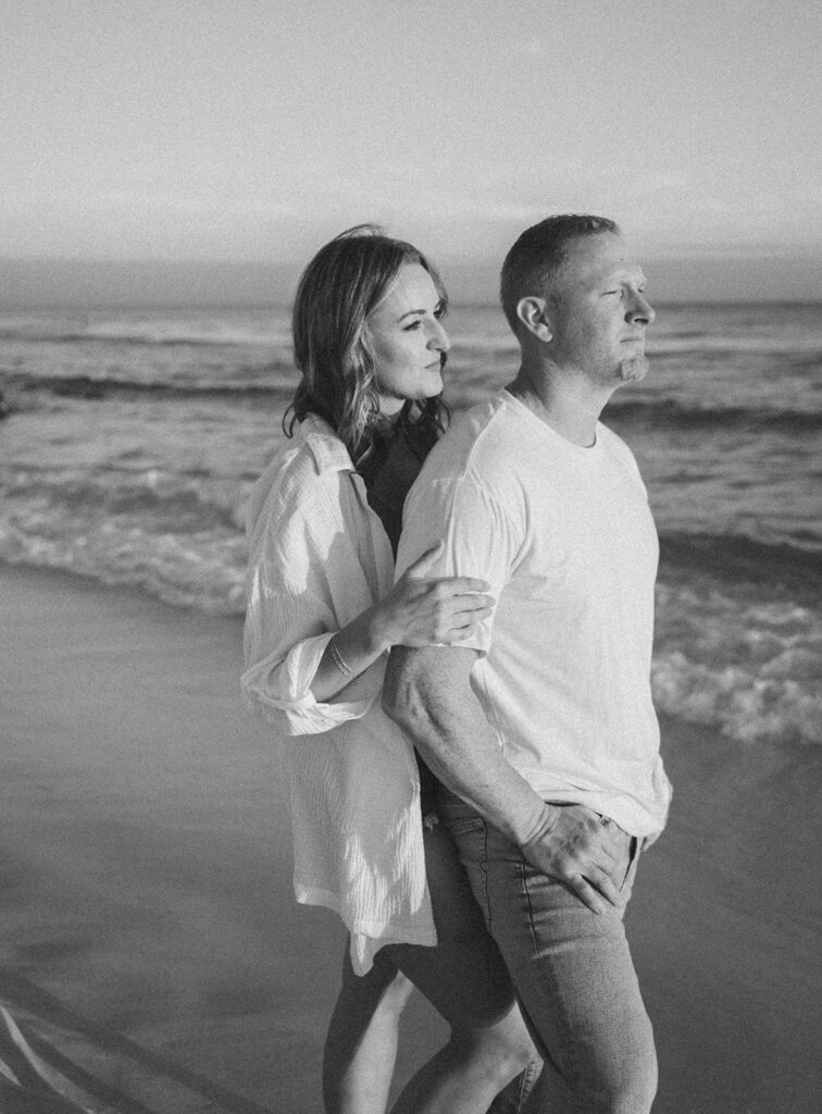 A woman stands behind a man on a beach, resting her hand on his arm. Both look out toward the ocean, appearing calm and thoughtful. This black and white image beautifully captures the serene mood of classic beach family pictures.