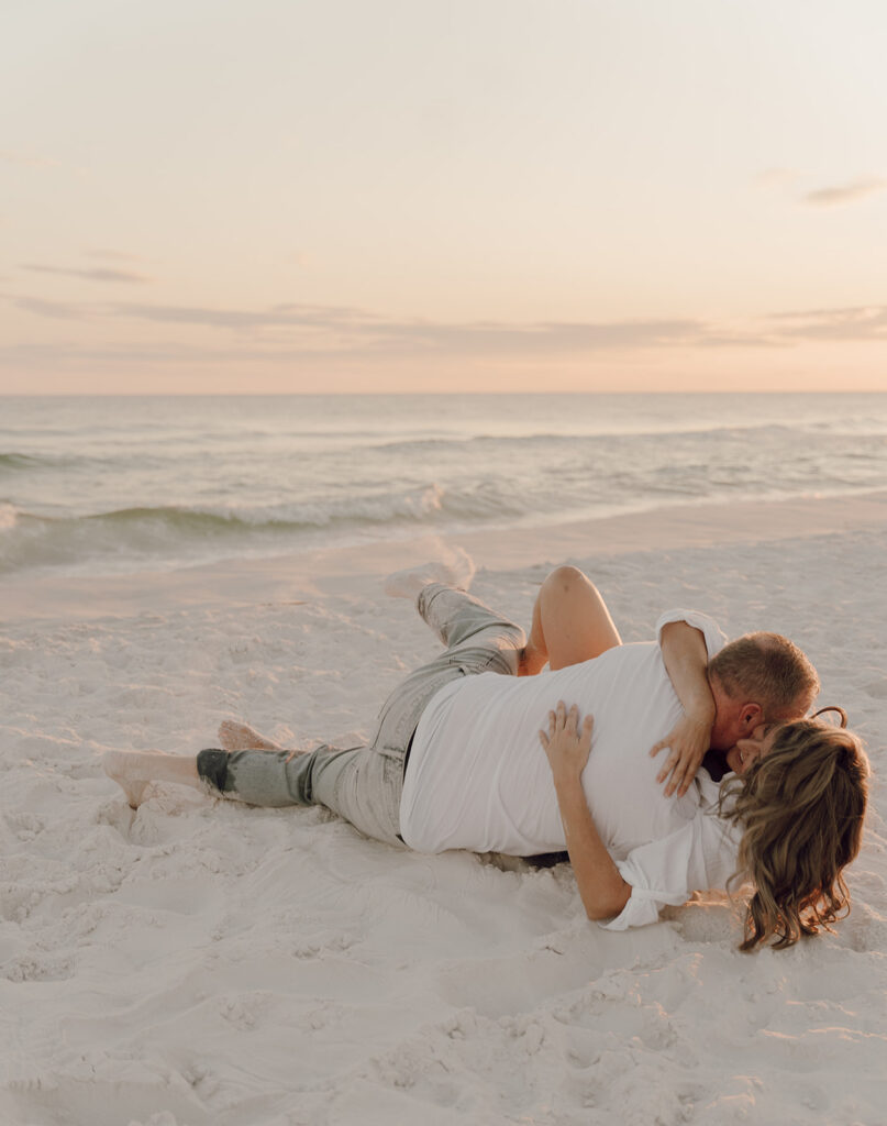 A couple lies on the sand at the beach, embracing as the sun sets over the calm ocean. The peaceful, romantic scene captures soft, warm light—perfect inspiration for seaside family pictures by a 30A family photographer.