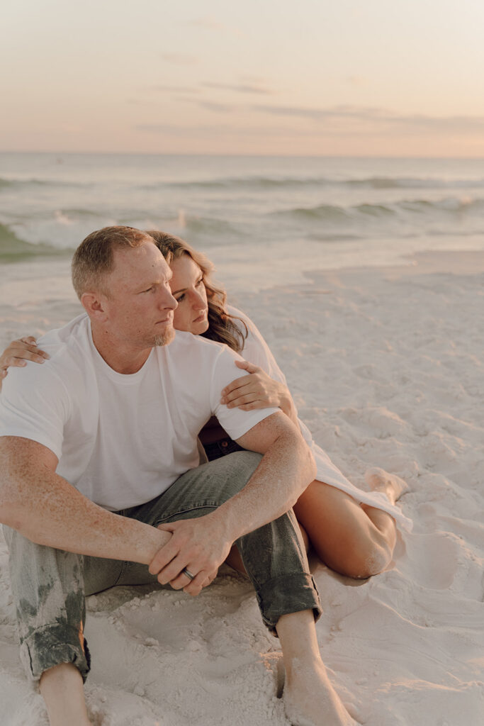 A couple sits close together on a sandy beach at sunset. The man in a white t-shirt and jeans looks thoughtful, while the woman hugs him from behind. This calm, intimate moment is perfect for beach family pictures.