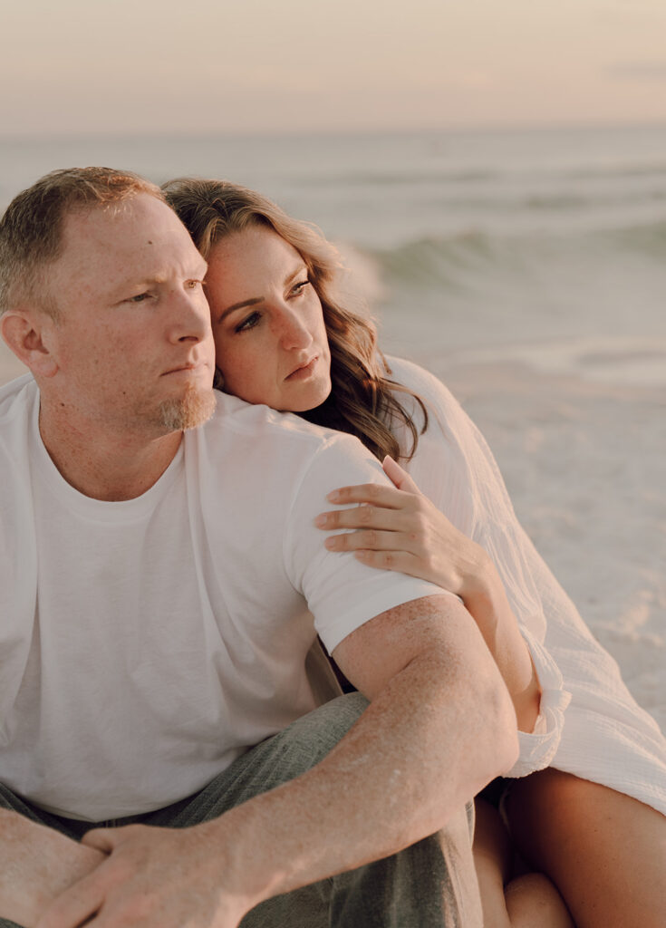 A couple sits on the beach at sunset, her head resting on his shoulder as they gaze into the distance. Wearing casual white tops by the shoreline, they create a tender moment reminiscent of documentary style family pictures.