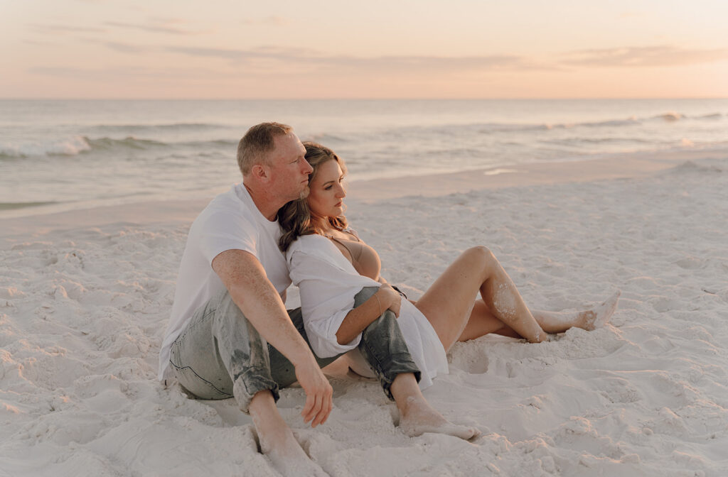A couple sits on white sand at the beach during sunset, leaning against each other and gazing at the ocean. Their light clothing and peaceful demeanor evoke the warmth often captured in documentary style family pictures.