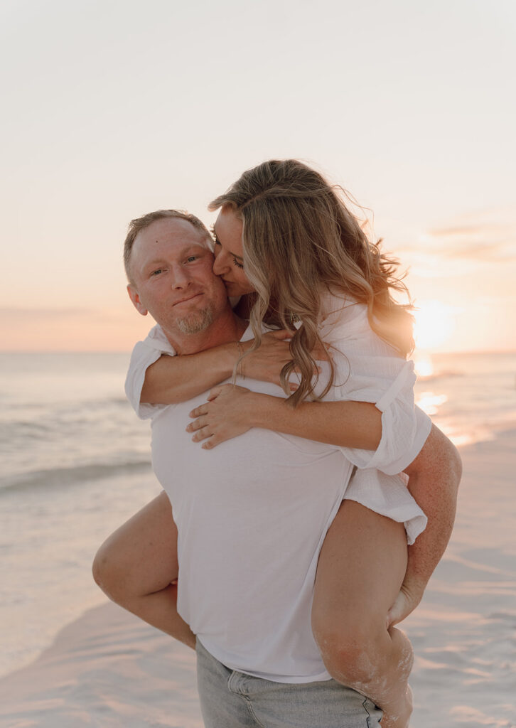 A man gives a woman a piggyback ride on a sandy beach at sunset, both smiling and wearing white shirts. The woman playfully nuzzles his cheek as the calm ocean glows behind them—perfect for those seeking Rosemary Beach family pictures.