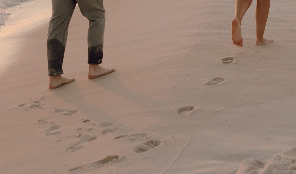 Two people walk barefoot along the sandy beach, leaving footprints in the soft sand as gentle waves approach the shore—a perfect moment for documentary style family pictures, showing only their legs and feet.