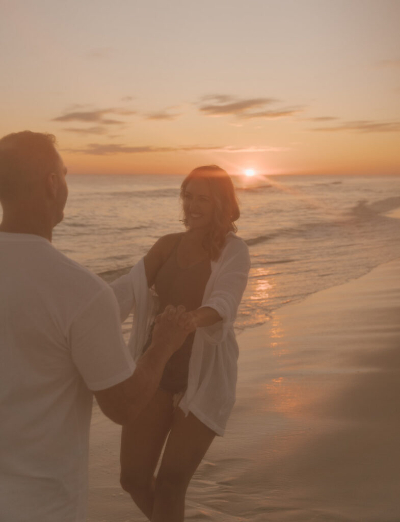 A couple holding hands and smiling at each other while standing on a beach at sunset, perfect for seaside family pictures, with gentle waves and an orange sky in the background.