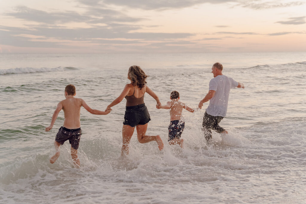 A family of four, holding hands, runs into gentle ocean waves at sunset, creating splashes as they enjoy the water together—perfect for candid seaside family pictures that capture real moments.