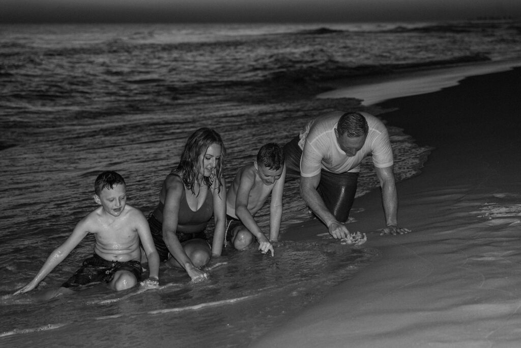 A black-and-white photo of two boys and two adults playing at the shoreline. They kneel in the shallow water, smiling as waves wash over the sand—capturing a joyful moment perfect for seaside family pictures.