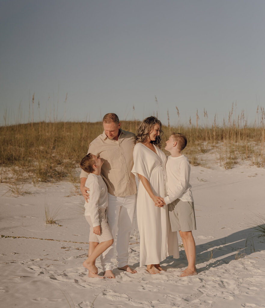 A family of four stands barefoot on a sandy beach, smiling and holding hands. Captured by a 30A family photographer, the parents and two young boys wear light, neutral clothing, with tall grass and blue sky as the perfect backdrop for beach family pictures.