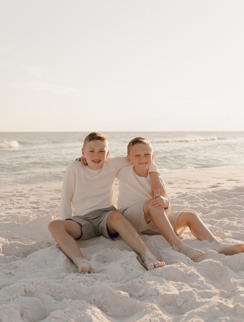 Two boys sit close together on a sandy shore by the ocean, both smiling. Wearing light shirts and shorts, one boy’s arm around the other, they enjoy warm, soft light—perfect for capturing candid beach family pictures at sunset.