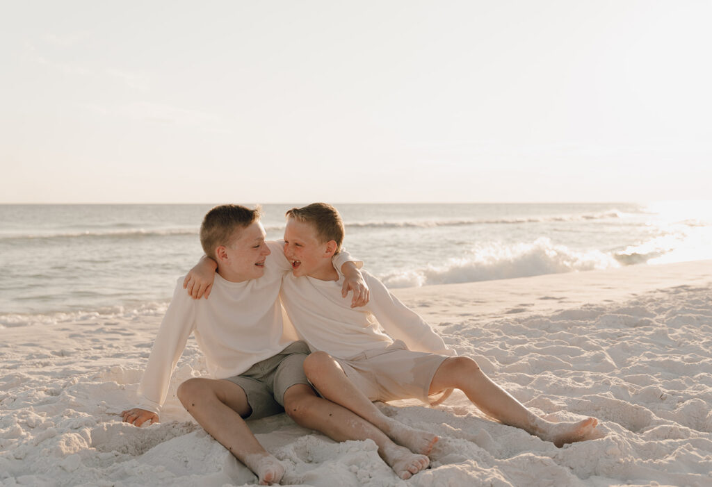 Two boys sit on white sand at the beach, smiling and laughing with their arms around each other. The sun sets over the calm ocean, casting a golden glow—perfect for capturing documentary style family pictures.