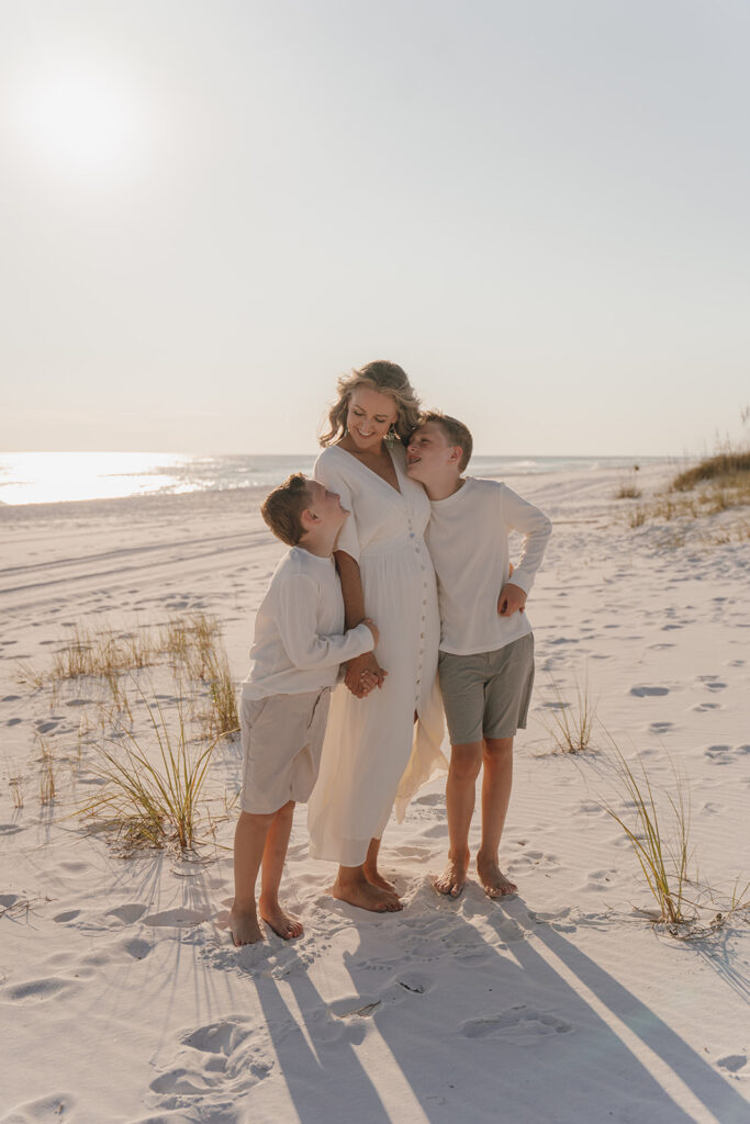 A woman in a white dress stands on a sandy beach at sunset, smiling while holding hands with two young boys. Grasses and footprints dot the sand—perfect for documentary style family pictures capturing the warmth of this seaside moment.