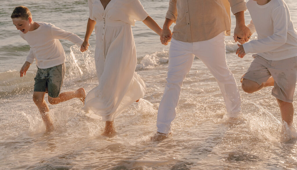 Four people, dressed in light-colored clothing, hold hands and walk through shallow water at the beach, waves splashing around their legs—a perfect moment for 30A family photographer capturing stunning beach family pictures. Photo shows only their lower bodies.