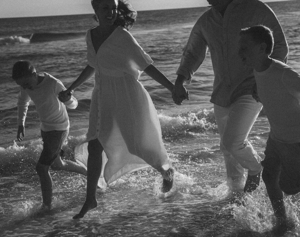 A family of four, dressed in light clothing, joyfully runs through shallow ocean waves at the beach, holding hands. This black and white image captures their happy expressions and movement—perfect for documentary style family pictures.
