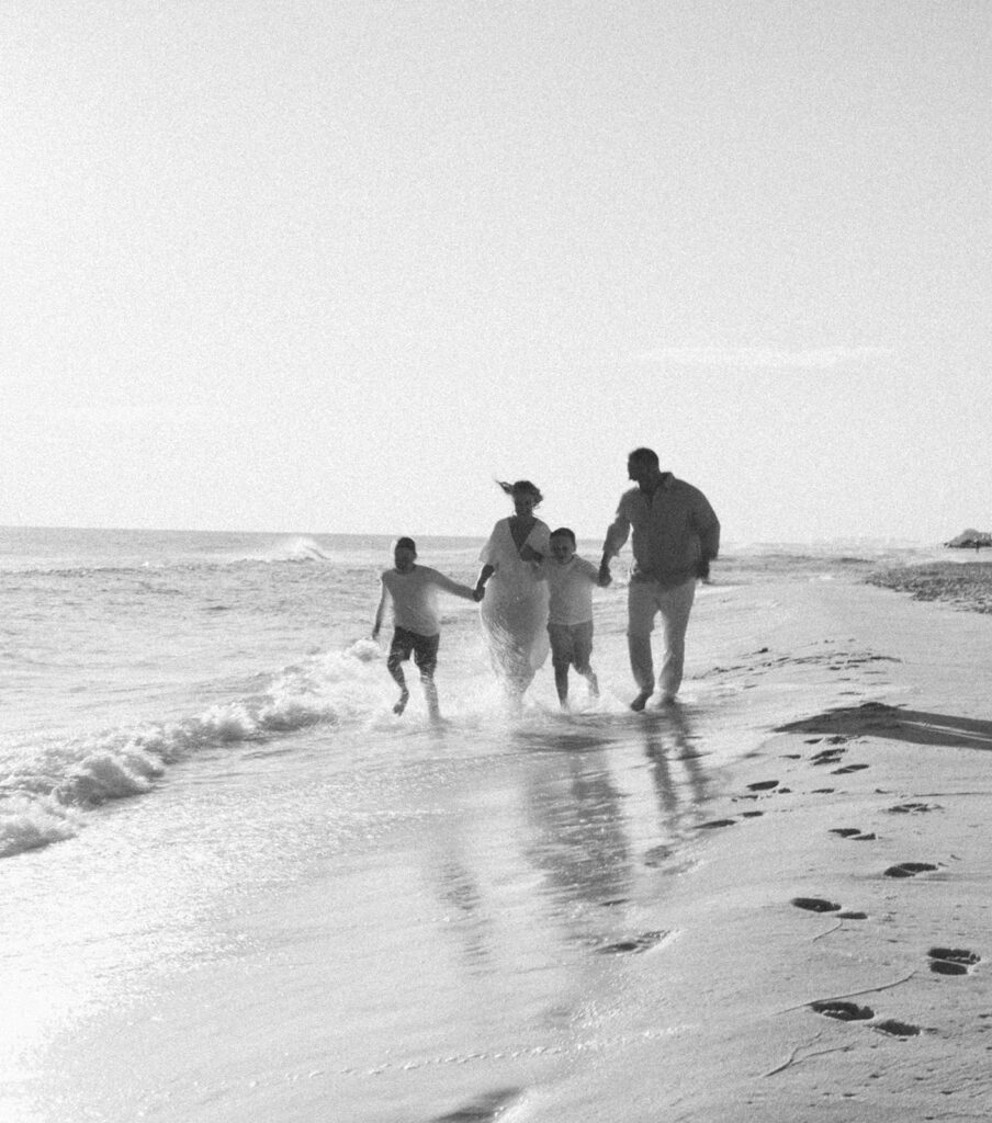 A family of four walks along the shoreline, holding hands and smiling, as gentle waves touch their feet on a bright, sunny day—perfect for timeless seaside family pictures with their reflections shining on the wet sand.