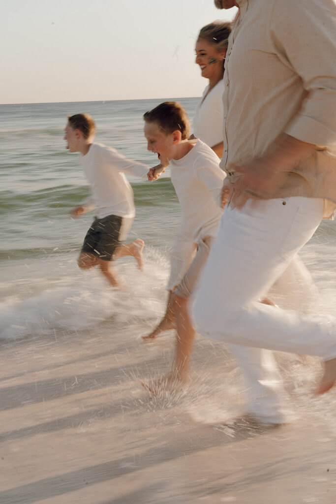 Four people, two adults and two children, run joyfully along the shoreline with waves splashing around their feet. Perfect for a 30A family photographer capturing documentary style family pictures full of laughter and genuine connection.