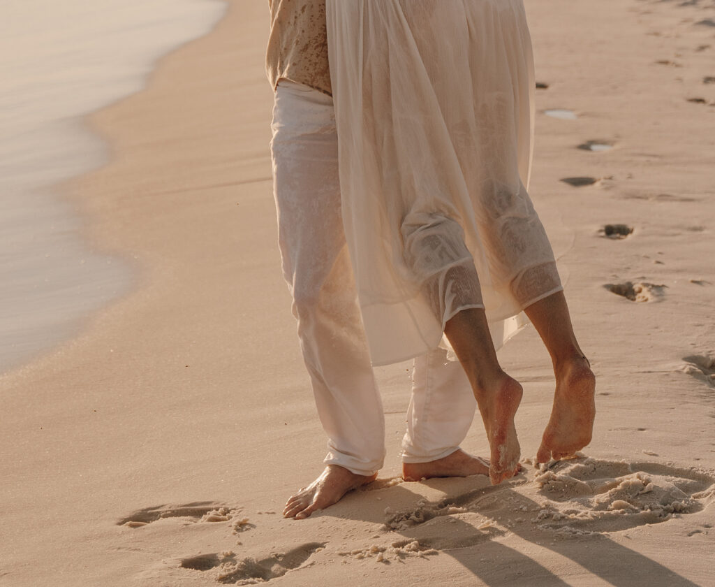 Barefoot couple walking closely together on a sandy beach near the water, leaving footprints behind. Their light, flowing clothing and the calm, intimate setting evoke the feel of documentary style family pictures.