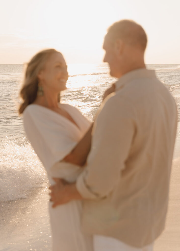 A softly blurred photo of a couple standing close together on the shore at sunset, smiling at each other with ocean waves and sunlight in the background—perfect for capturing authentic, documentary style family pictures.