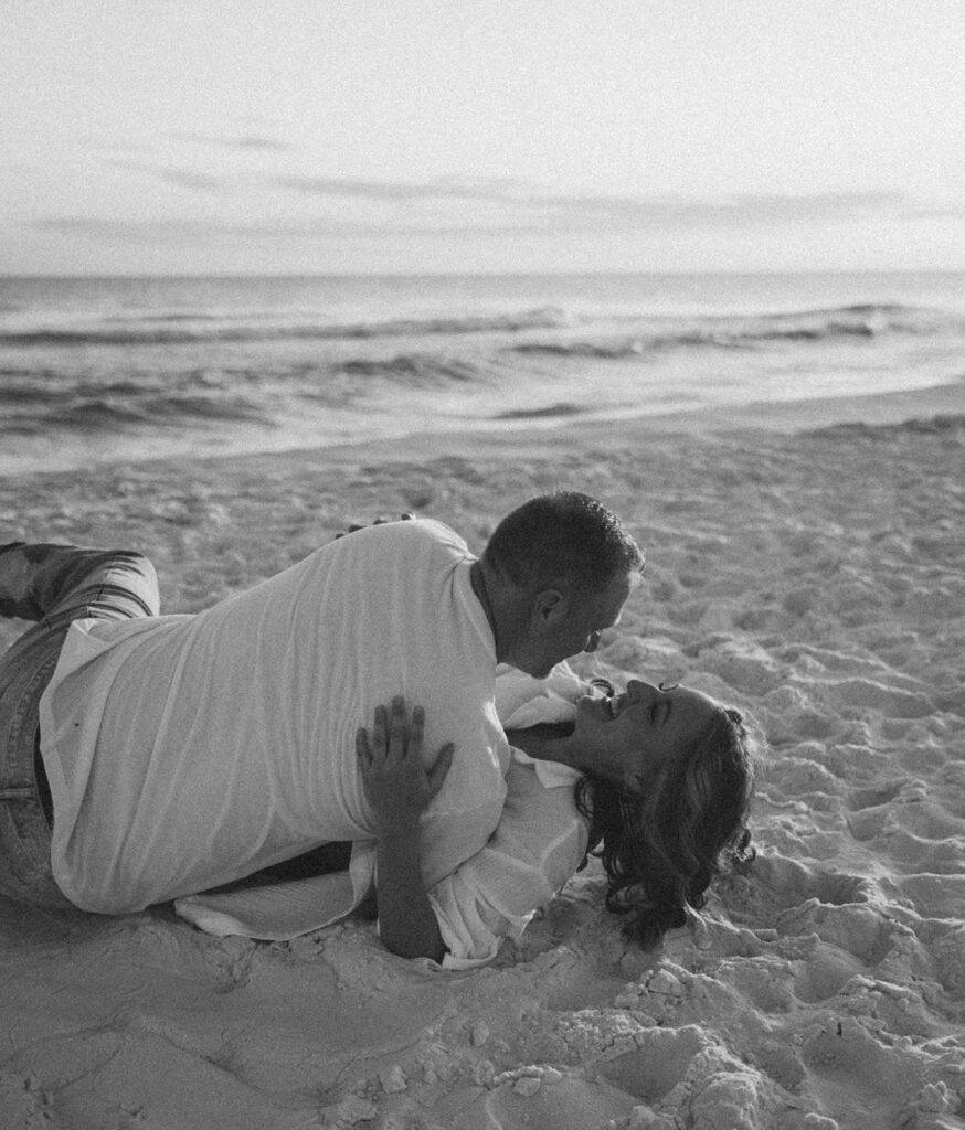 A couple lies on the sand at the beach, laughing and embracing under a bright sky, gentle waves behind them—a timeless black and white shot reminiscent of documentary style family pictures.