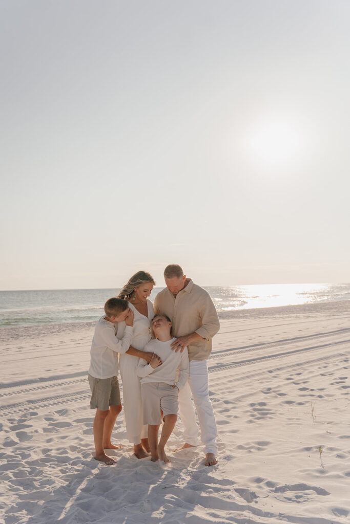 A family of four stands closely together on a sandy beach at sunset. The parents and two young boys, dressed in light neutrals, embrace warmly near the shoreline—an ideal moment for seaside family pictures by a 30A family photographer.