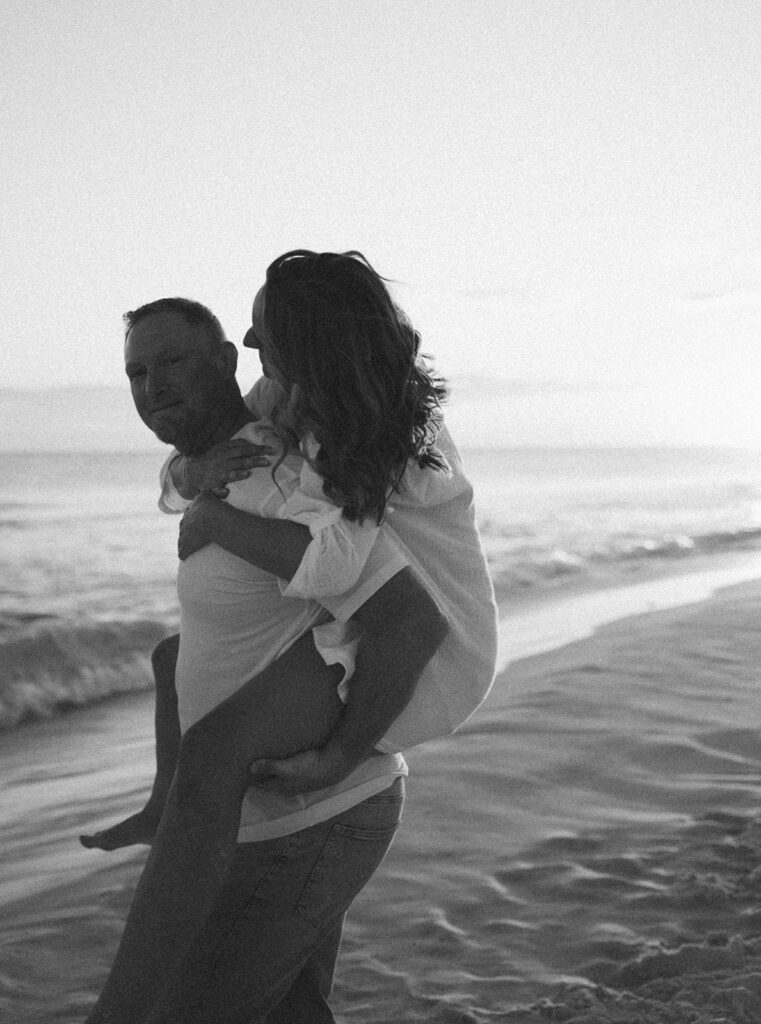 A man carries a woman on his back along a sandy shore, both smiling in light clothing. This black and white moment captures the joy found in beach family pictures with gentle ocean waves rolling in behind them.