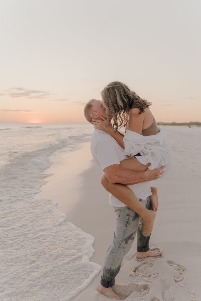 A man stands in shallow ocean waves at sunset, holding a woman wrapped in a white shirt as they kiss. Both are barefoot on the sandy beach, evoking the warmth and intimacy often found in documentary style family pictures.