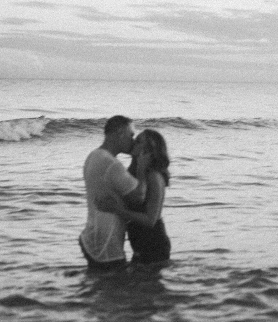 A couple stands in the ocean, embracing and kissing in shallow water. The image is black and white and slightly out of focus, with gentle waves in the background—perfect inspiration for dreamy seaside family pictures.