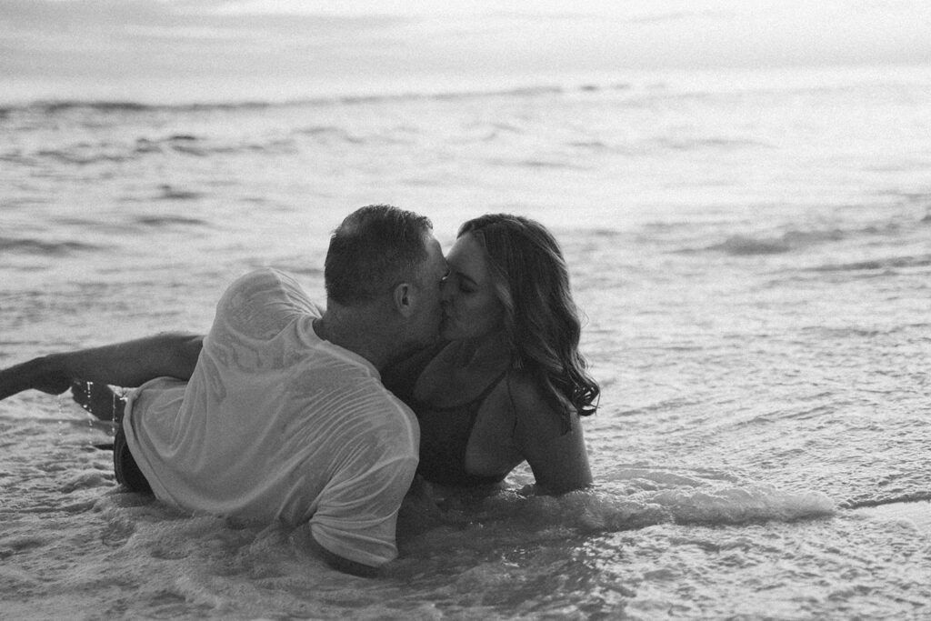 A couple lies in shallow ocean water, kissing and embracing with gentle waves around them. This black and white image, captured by a 30A family photographer, creates a romantic and intimate atmosphere.
