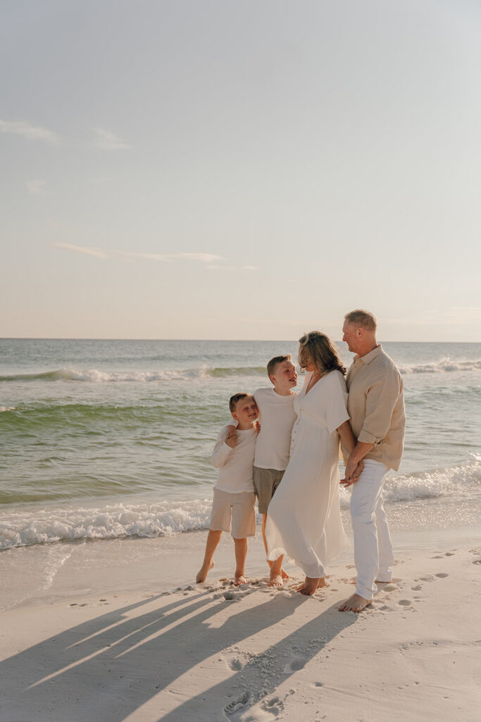 A family of four stands barefoot on the beach near the shoreline, smiling and embracing as gentle waves roll in under a clear sky during sunset—perfect for capturing candid, documentary style family pictures.