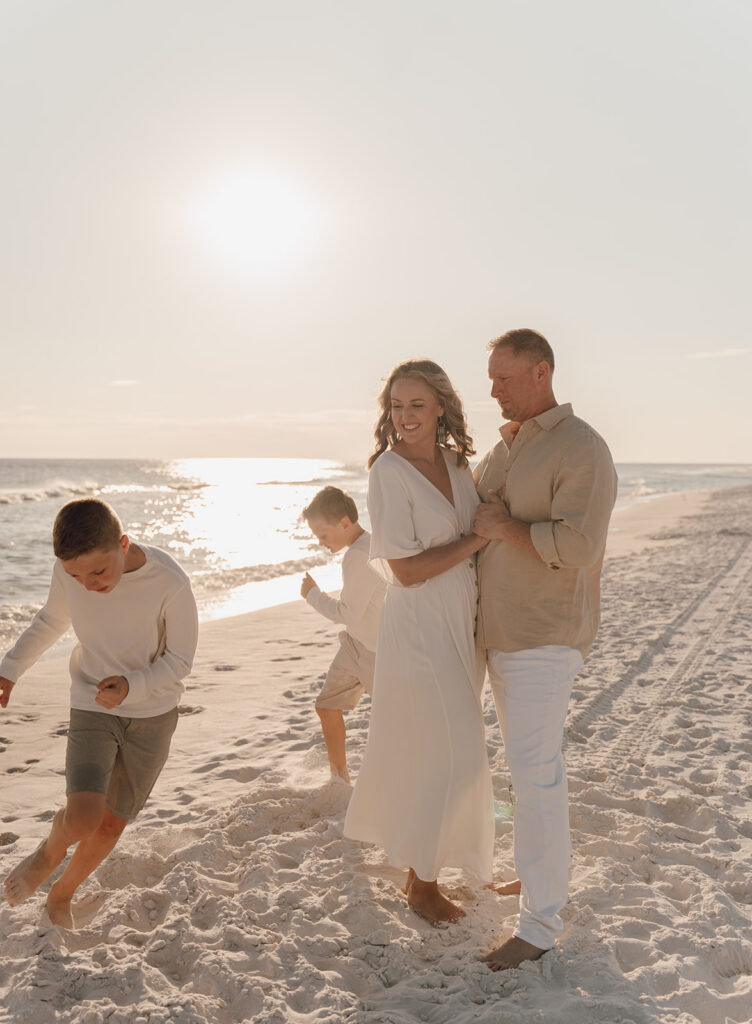 A family of four enjoys a sunny day by the ocean, captured in beautiful beach family pictures. The parents smile and embrace while the children play in the sand, creating memories along the shoreline under the shining sun.