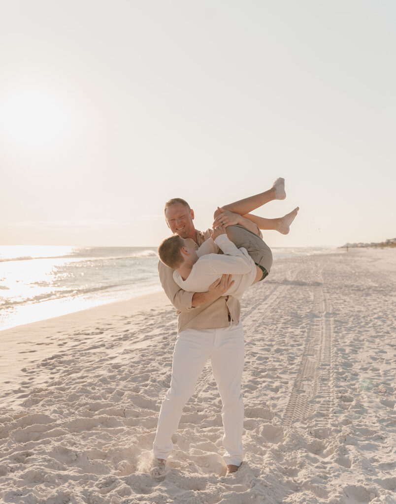 Two men smile as one playfully lifts and holds the other sideways on a sandy beach at sunset, capturing the joy of seaside family pictures with gentle waves and tire tracks in the background.