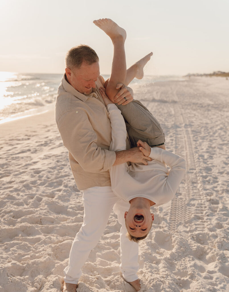 A man in light clothing playfully holds a laughing child upside down on a sandy beach near the ocean, perfect for candid seaside family pictures with tire tracks and waves visible in the background.