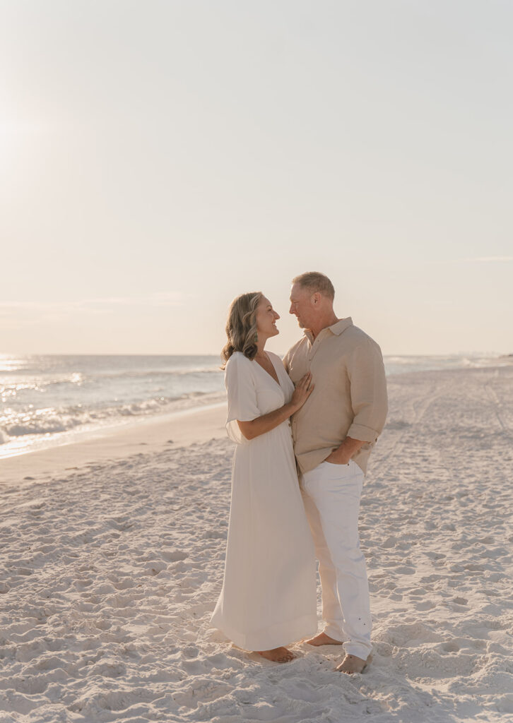 A couple stands barefoot on a sandy beach at sunset, facing each other and smiling. The woman wears a white dress and the man wears a beige shirt and white pants—perfect for relaxed, documentary style family pictures by the ocean.