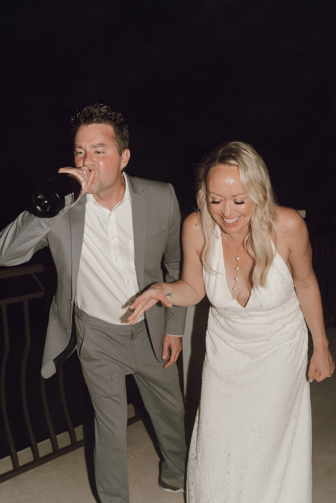 Groom drinking champagne out of the bottle while the bride laughs. 