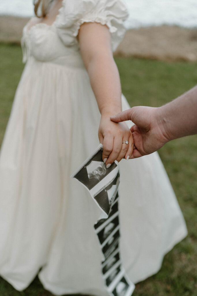A woman in a white flowy dress holding an ultrasound photo strip while her husband holds her hand. 