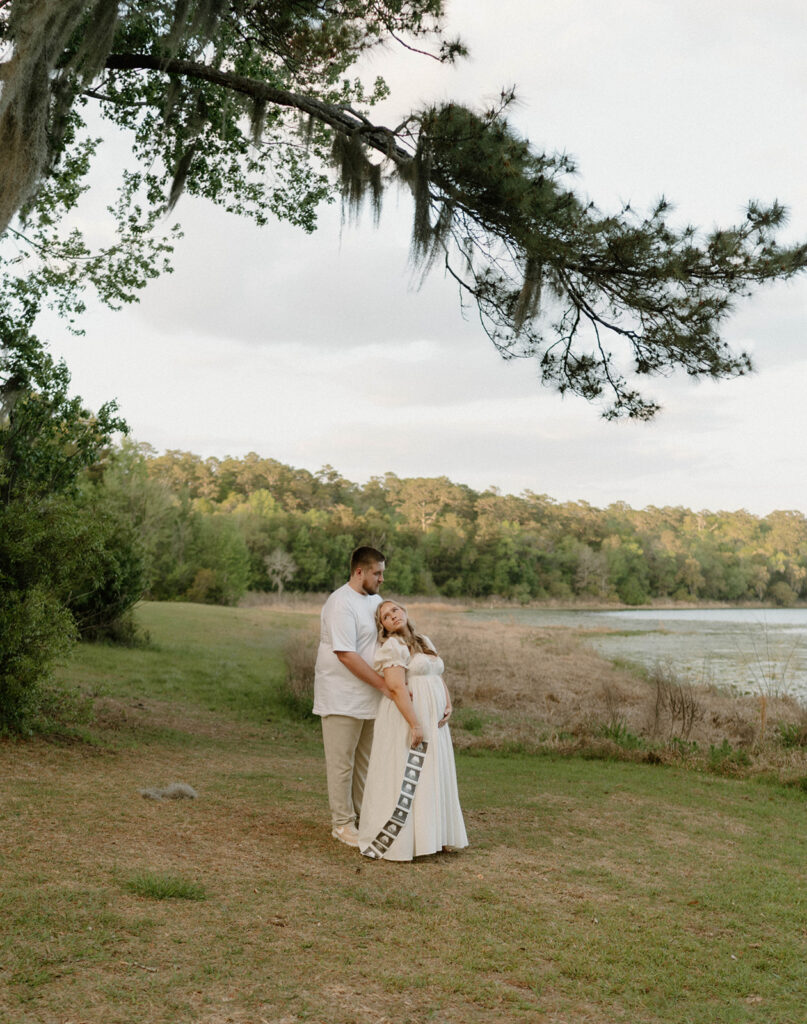 A man holding his wife by a lake for pregnancy announcement photos. 