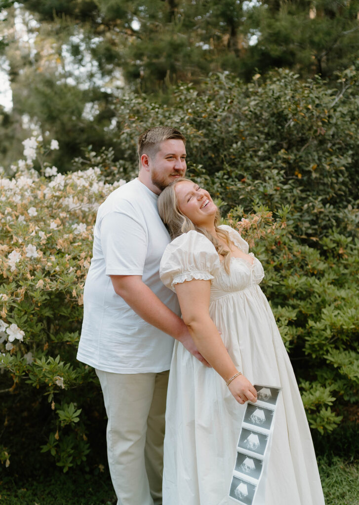 Couple smiling by a flower bush for pregnancy announcement photos.