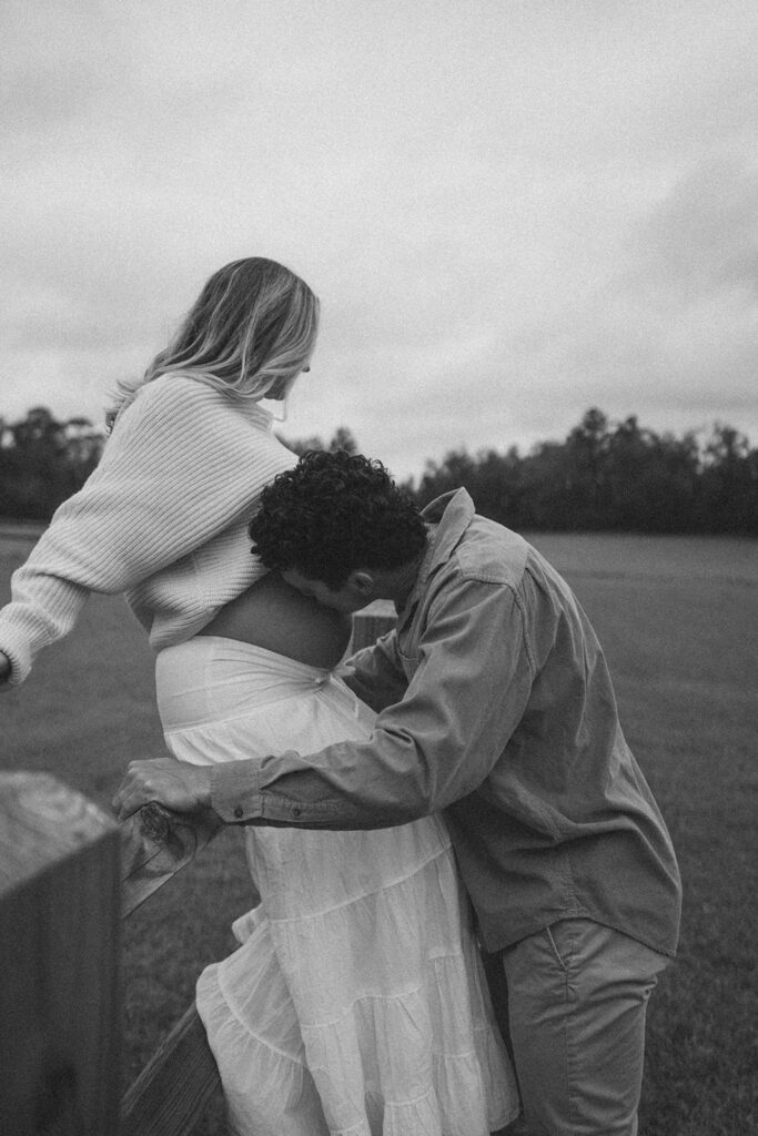 A woman sitting on a fence while her husband kisses her pregnant belly.
