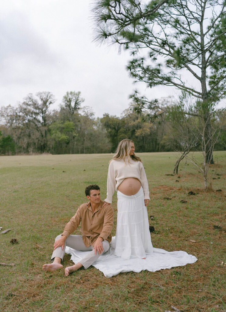 Maternity photos of a man sitting on a white blanket with his wife standing next to him.