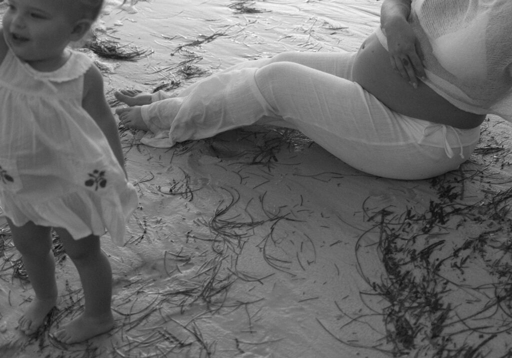 Toddler playing in the sand next to her mother.