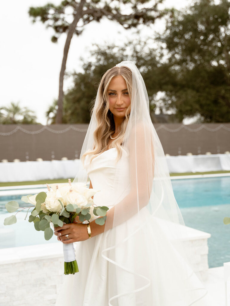 Bride standing by a pool, holding her bouquet.
